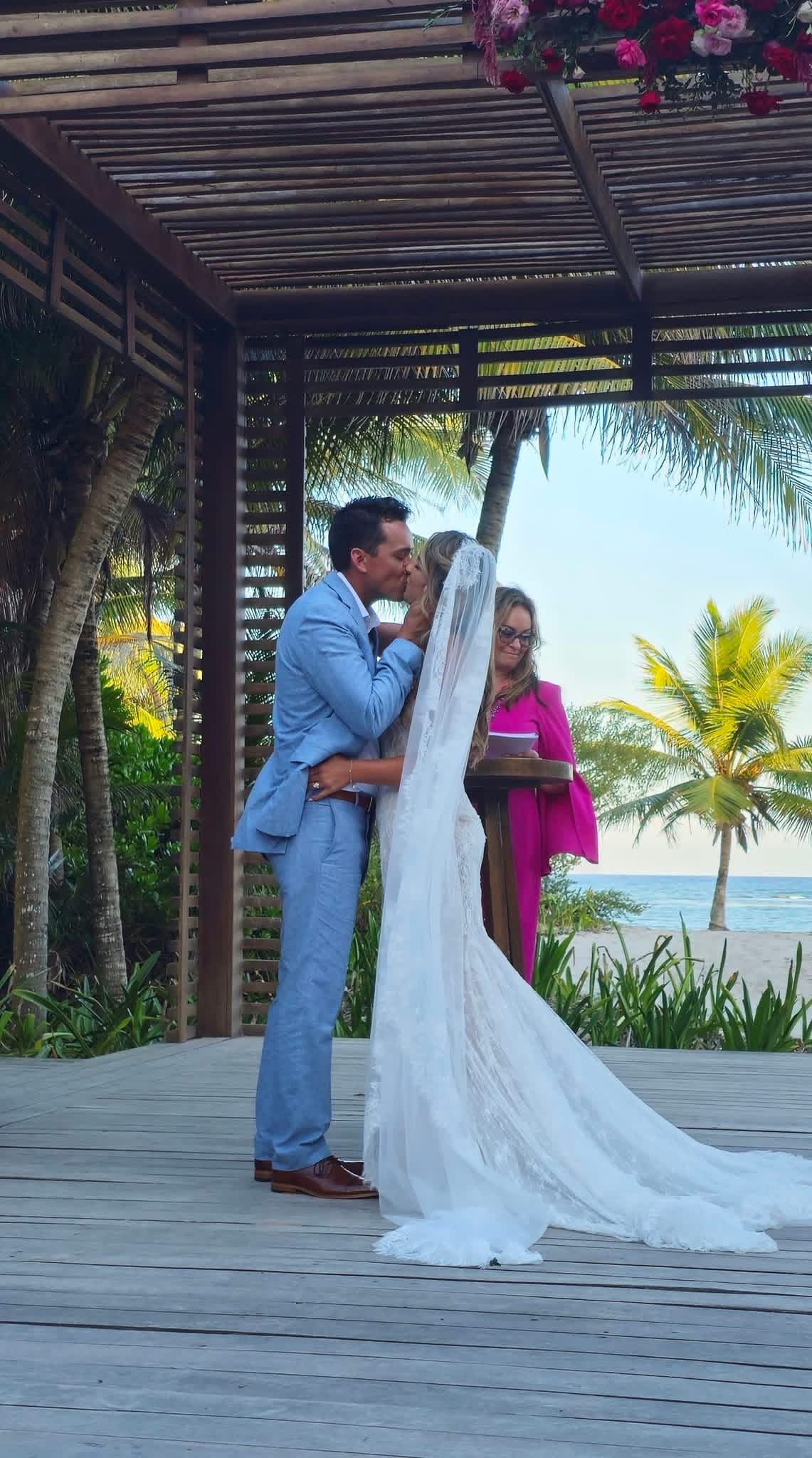 The newlyweds kissing with Amy behind them and the ocean in the background